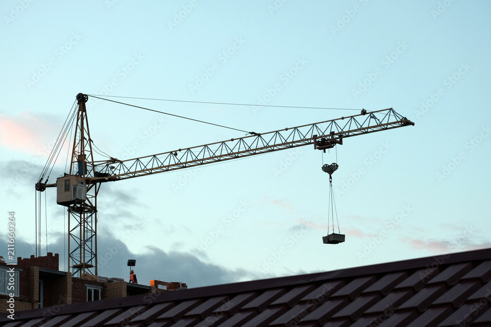Home construction. Arrow of a tower crane with a load against the background of the evening sky. Tiled roof in the foreground.