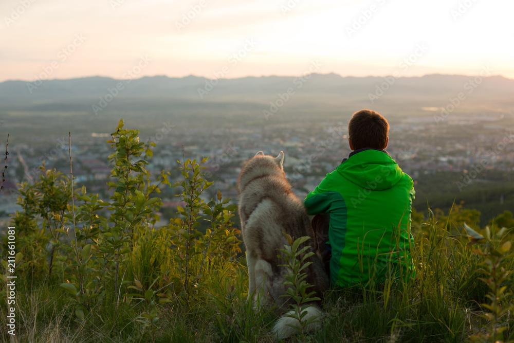 Image of a man in the green jacket with cute siberian husky dog sitting and looking at mountain view at sunset