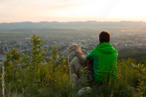 Image of a man in the green jacket with cute siberian husky dog sitting and looking at mountain view at sunset