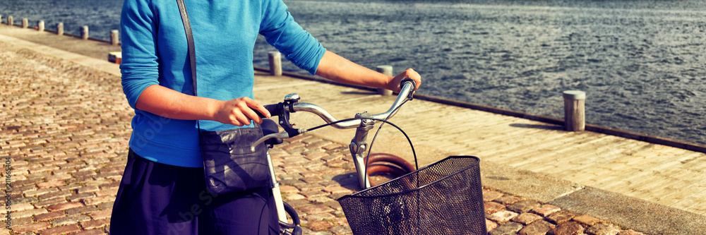 Obraz premium Bicycle commute in city street. Girl biking with bicycle walking on sidewalk in urban harbour old port. Woman in European city. Active healthy lifestyle. Banner panorama.