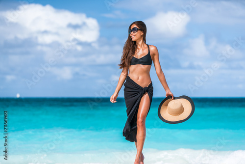 Beach vacation luxury travel elegant bikini woman in fashion swimwear sun tanning in tropical destination getaway. Asian girl wearing cover-up pareo, sunglasses, hat. Body skin care suntan protection.