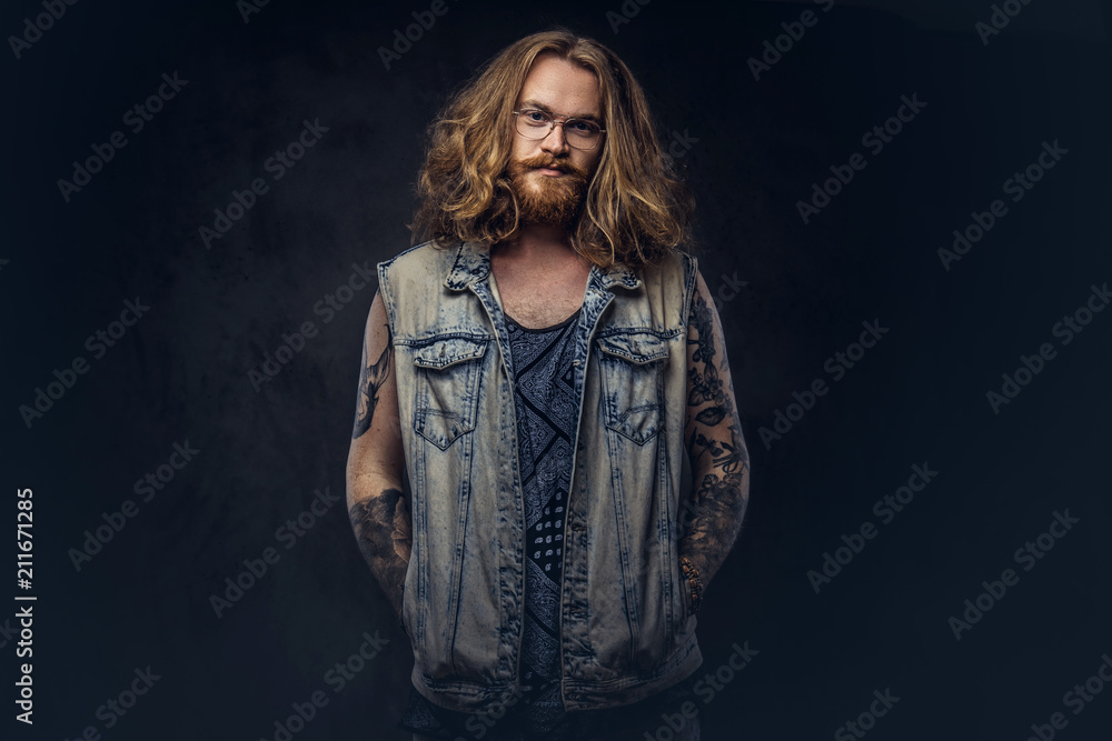 Redhead hipster male with long luxuriant hair and full beard dressed in casual clothes posing with hands in pockets in a studio, looking a camera. Isolated on a dark background. Redhead hipster male with long luxuriant hair and full beard dressed in casual clothes posing with hands in pockets in a studio, looking a camera. Isolated on a dark background.