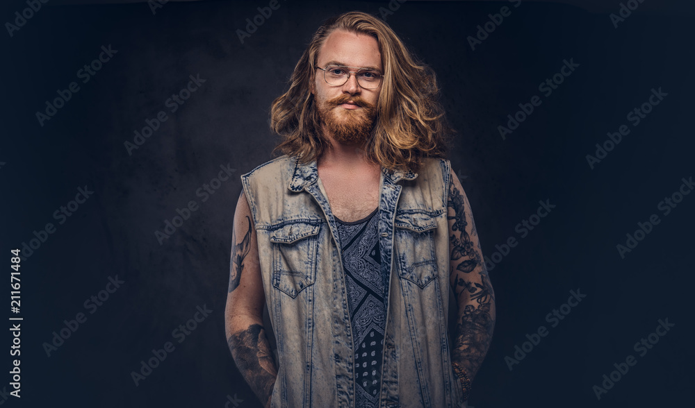 Redhead hipster male with long luxuriant hair and full beard dressed in casual clothes posing with hands in pockets in a studio, looking a camera. Isolated on a dark background. Redhead hipster male with long luxuriant hair and full beard dressed in casual clothes posing with hands in pockets in a studio, looking a camera. Isolated on a dark background.
