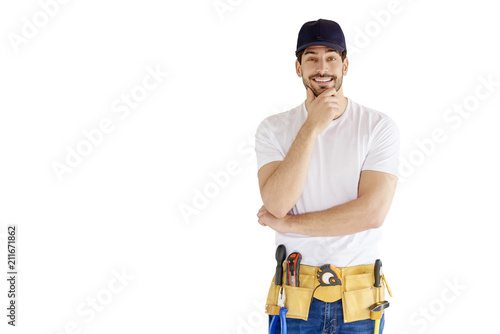 Portrait of young handyman standing at isolated white background with copy space. Successful repairman wearing baseball cap and tool belt.