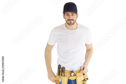 Portrait of young handyman standing at isolated white background with copy space. Successful repairman wearing baseball cap and tool belt. photo