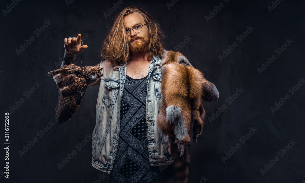 Portrait of tattoed redhead hipster male with long luxuriant hair and full beard dressed in a t-shirt and jacket holds a keeps the scarecrow of an owl and fox skin on a shoulder in a studio. Portrait of tattoed redhead hipster male with long luxuriant hair and full beard dressed in a t-shirt and jacket holds a keeps the scarecrow of an owl and fox skin on a shoulder in a studio.