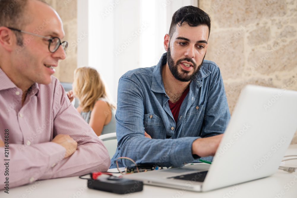 Two men sitting in front of a laptop discussing Two men sitting in front of a laptop discussing