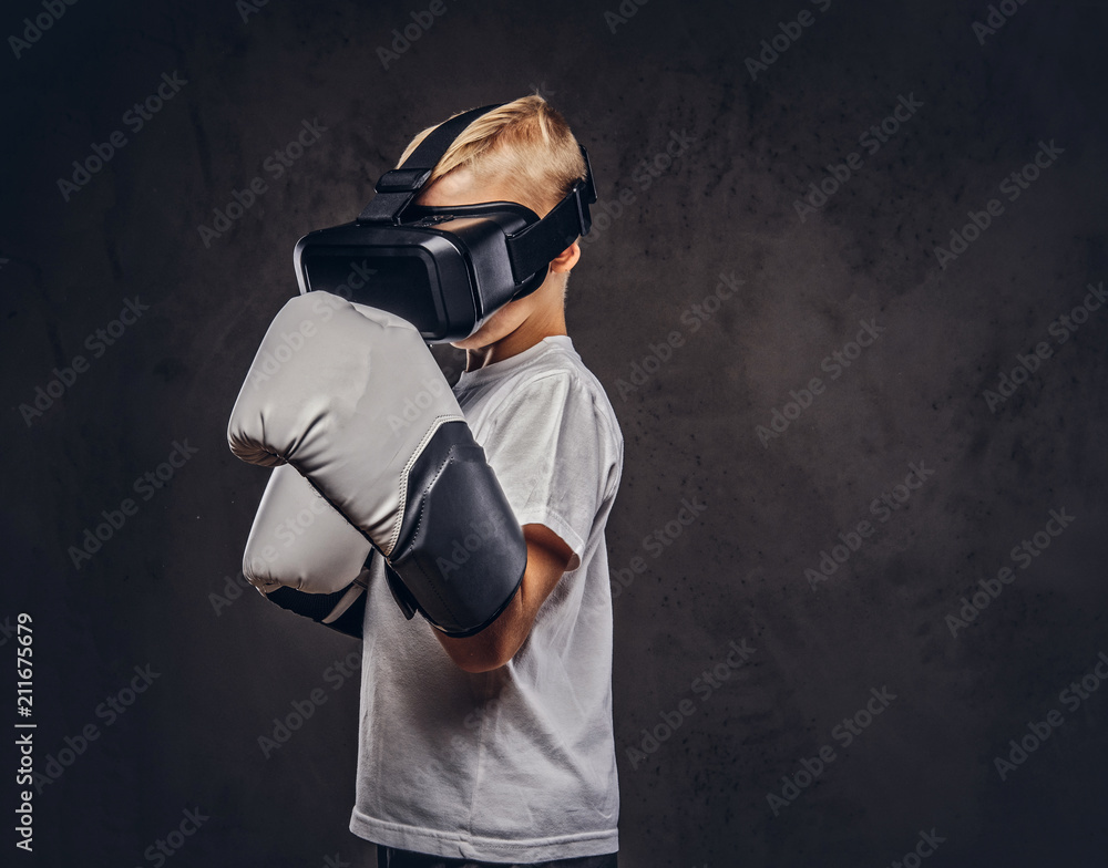 Schoolboy boxer with blonde hair dressed in a white t-shirt wearing visual reality glasses and boxing gloves, workout in a studio. Isolated on dark textured background.