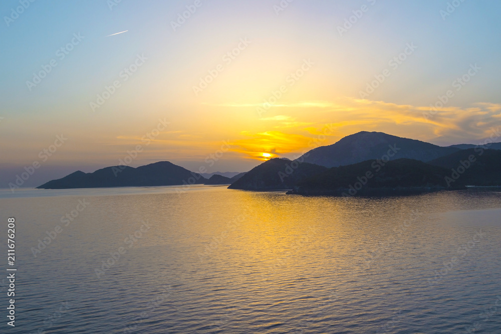 Mediterranean landscape. Sunset on the sea with flowing bright colored rays of the sun through the clouds. Silhouettes of mountains. Silhouettes the ship. Background.