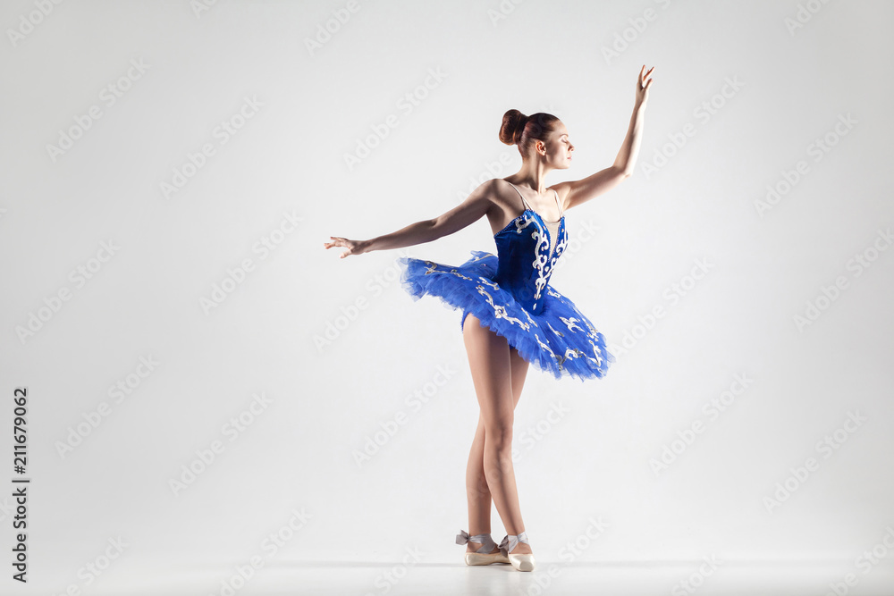 attractive ballerina with bun collected hair wearing blue dress and pointe shoes performing in white studio. attractive ballerina with bun collected hair wearing blue dress and pointe shoes performing in white studio.