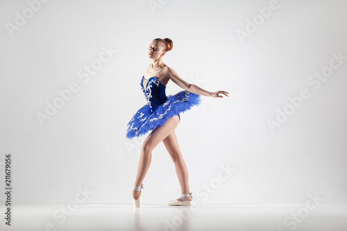 attractive ballerina with bun collected hair wearing blue dress and pointe shoes performing in white studio.