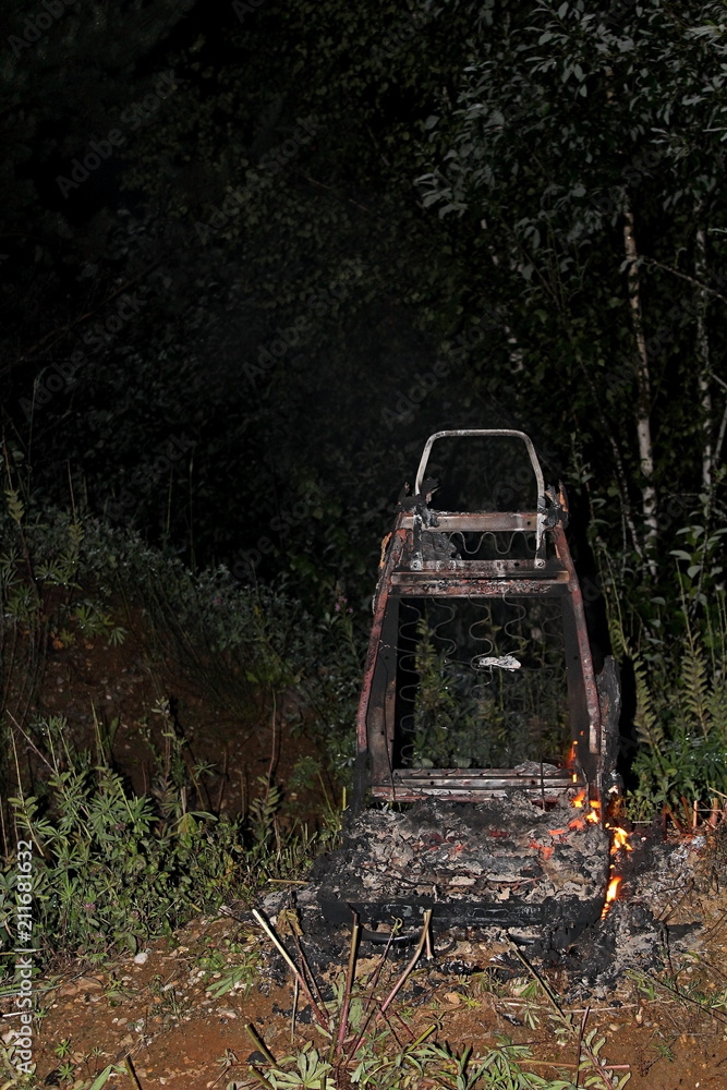 Automobile seat, covered in flames, in the open air, on a dark summer night.