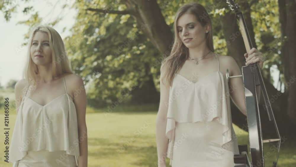 Two girls in dresses are walking along the lawn holding a violin and a cello. Two girls in dresses are walking along the lawn holding a violin and a cello.