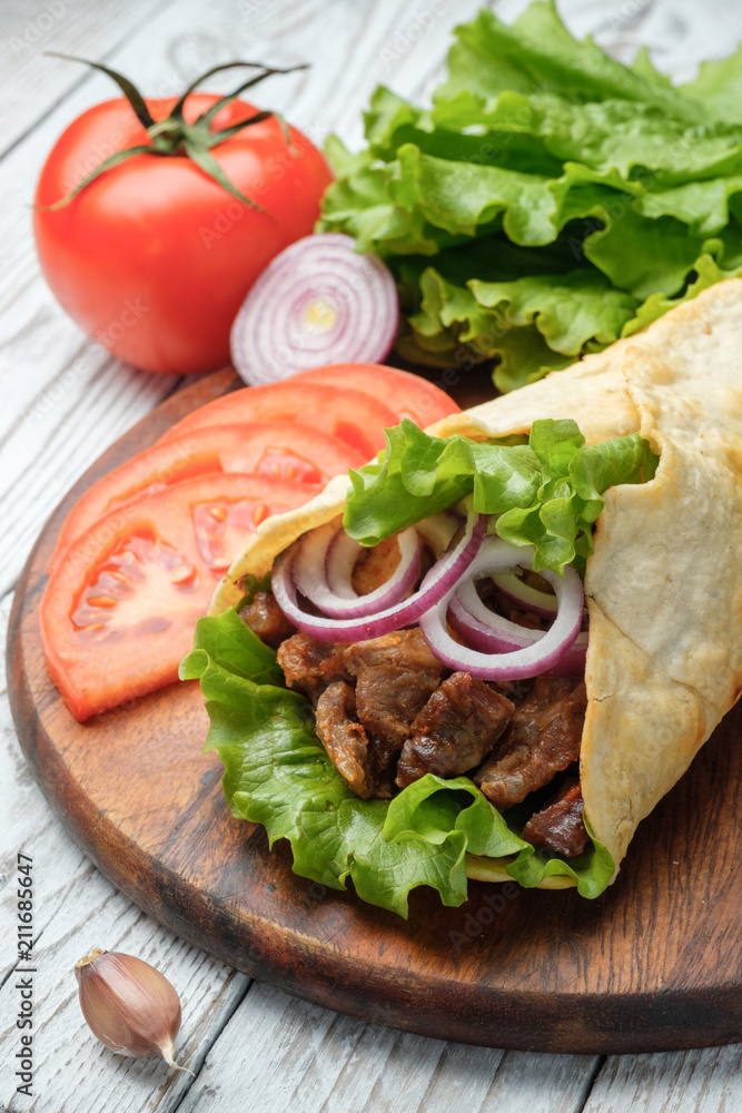 Doner kebab is lying on the cutting board. Shawarma with meat, onions, salad lies on a white old wooden table. Doner kebab is lying on the cutting board. Shawarma with meat, onions, salad lies on a white old wooden table.