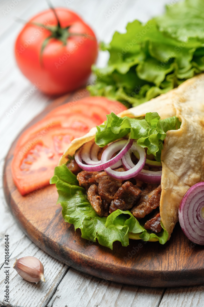 Doner kebab is lying on the cutting board. Shawarma with meat, onions, salad lies on a white old wooden table. Doner kebab is lying on the cutting board. Shawarma with meat, onions, salad lies on a white old wooden table.