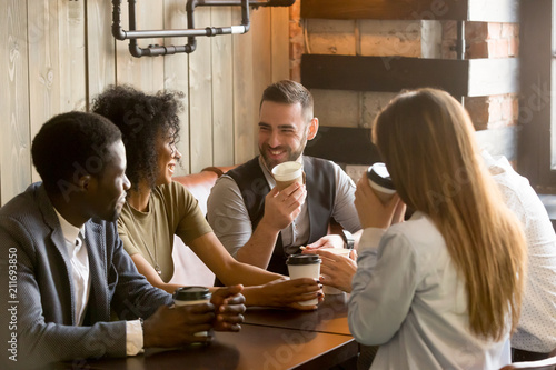 Smiling multiracial people enjoying coffee, talking spending pleasant time together in cozy coffee shop, diverse friends drinking from paper cups, meeting at work break for casual conversation in cafe