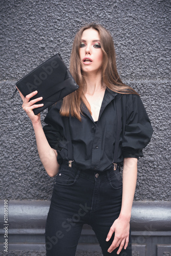 Beautiful girl with a black clutch in her hand. Woman in dark clothes on a dark background in the street. Toned image.