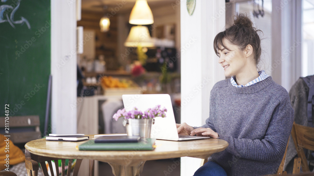 A young smiling delightful cute happy brunette girl dressed in a grey pullover is working with a laptop in a cafe