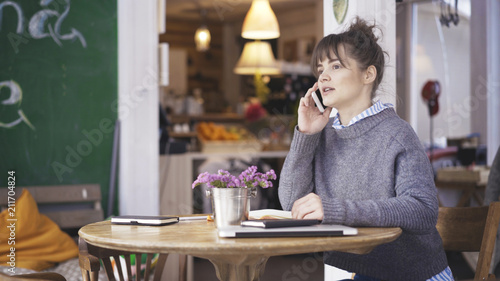 A young surprised cute happy brunette girl dressed in a grey pullover is having a call in a cafe
