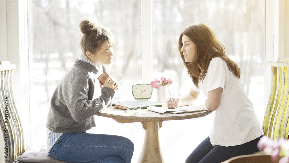 Two female friends busy sitting in a cafe face to face preparing for the exam talking making notes Two female friends busy sitting in a cafe face to face preparing for the exam talking making notes