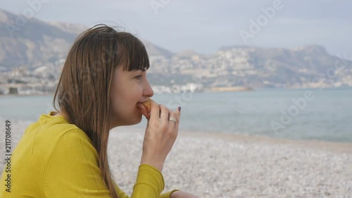 Portrait of young female eating at the seaside. Attractive caucasian woman biting sandwich on the off-season beach