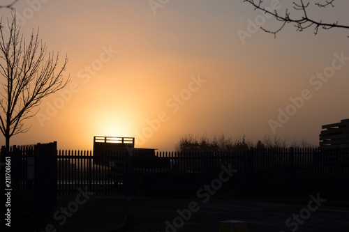 Orange misty sun behind trailer and metal fencing in an industrial estate