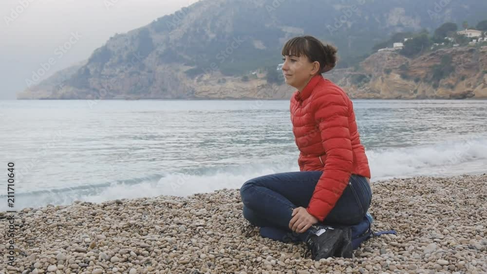 Young female on sea background. Attractive caucasian woman in red bubble jacket and blue jeans sitting on a pebble beach on sea background. Girl smiling peacefully looking forward Young female on sea background. Attractive caucasian woman in red bubble jacket and blue jeans sitting on a pebble beach on sea background. Girl smiling peacefully looking forward