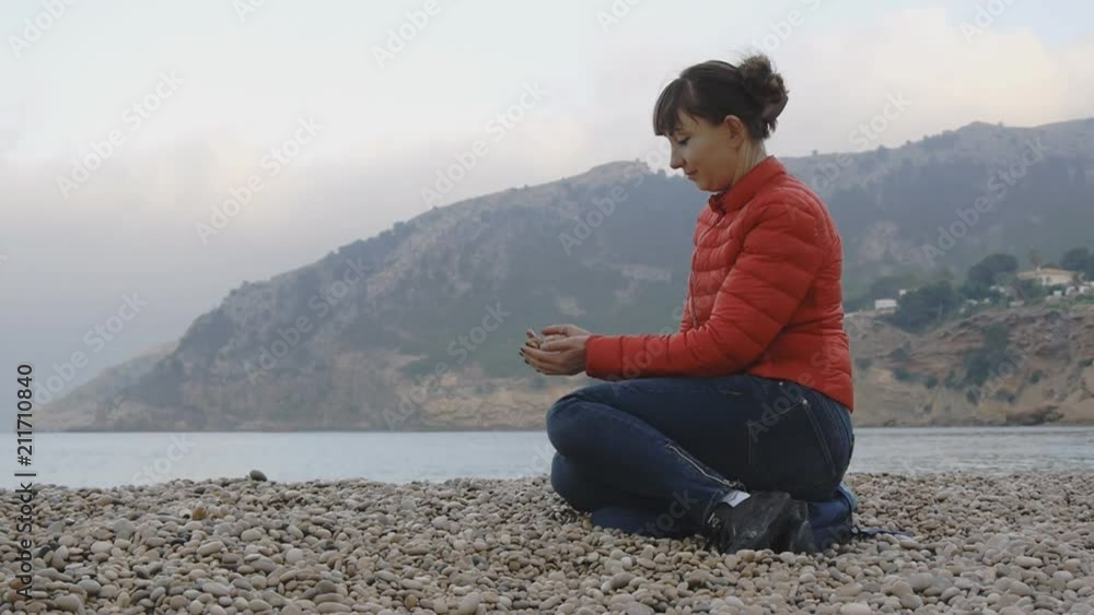 Young female on sea background. Attractive caucasian woman in red bubble jacket and blue jeans sitting on a pebble beach on sea background. Girl touches the stones from the palm to the palm Young female on sea background. Attractive caucasian woman in red bubble jacket and blue jeans sitting on a pebble beach on sea background. Girl touches the stones from the palm to the palm