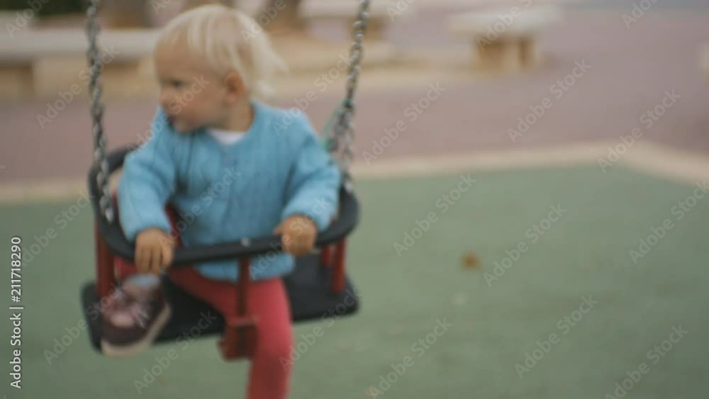 Baby in playground. Naughty blonde baby girl in blue knitted jersey riding a chain swing Baby in playground. Naughty blonde baby girl in blue knitted jersey riding a chain swing