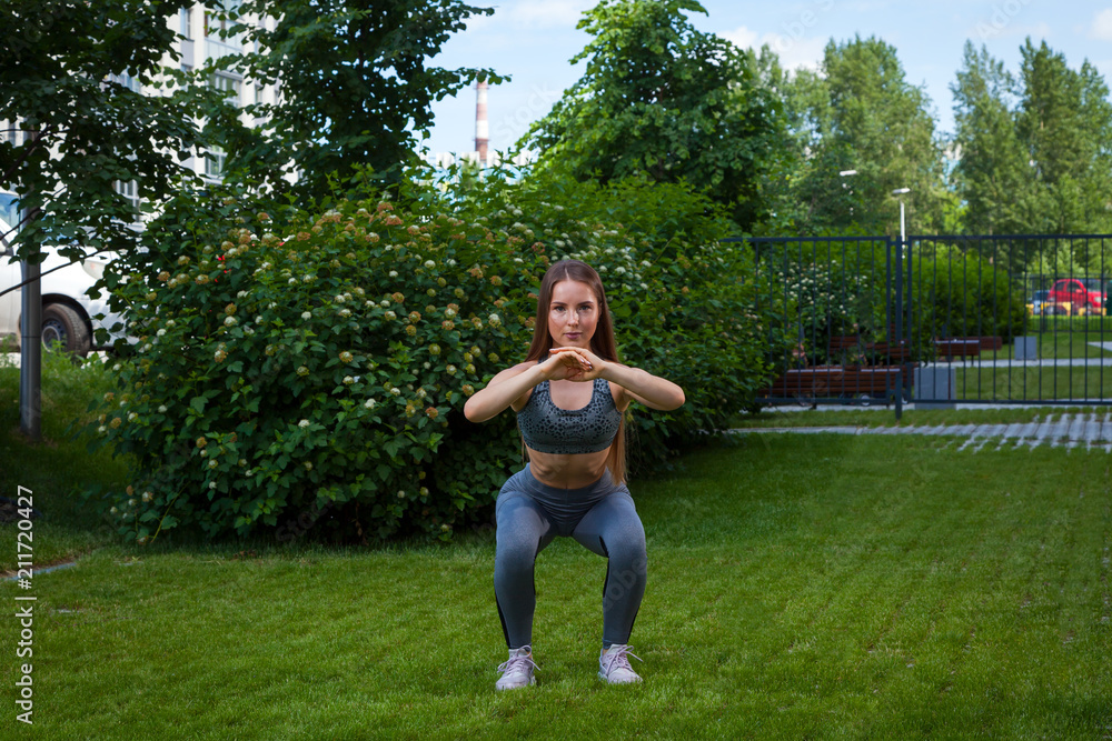 A dark-haired woman coach in a sporty short top and gym leggings shows the correct squatting technique, the position of a full squat, hands in front, on a summer day in a park on a green lawn