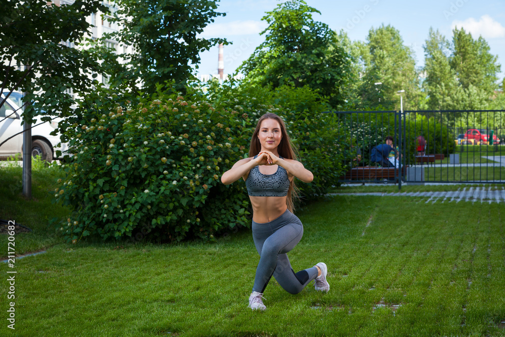 A  trainer in a sporty short top and gym leggings shows the correct technique of lunging the legs to inflate the buttocks, the position of a full squat, hands in front on a summer day 