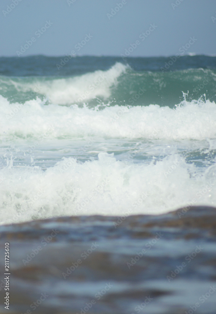 Fototapeta premium The foam of waves breaking over a brown rock. The water is pale aqua, becoming darker towards the horizon.