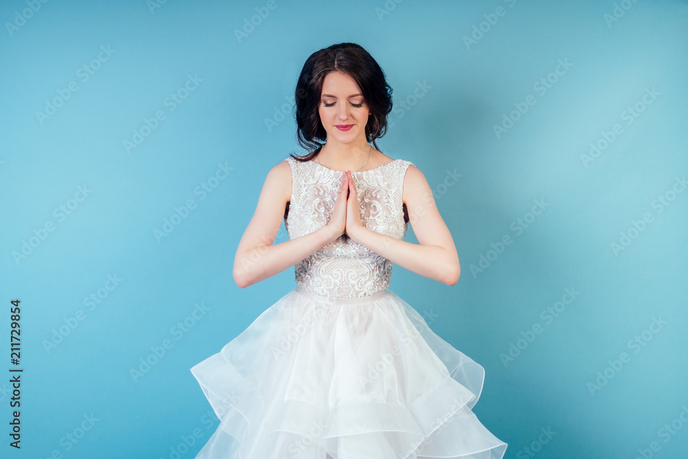 young and beautiful calm woman bride with makeup in chic white wedding dress practicing yoga and meditating in studio on a blue background .