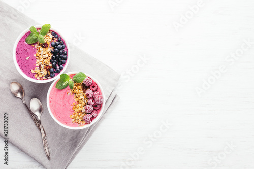 Two smoothie bowls with granola, blueberries, raspberries and mint on white wooden table. Healthy breakfast, top view.