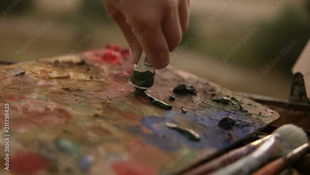 Close up of a woman's hand artist squeezes out the paints from the tube on palette. Preparing for art work outdoors Close up of a woman's hand artist squeezes out the paints from the tube on palette. Preparing for art work outdoors