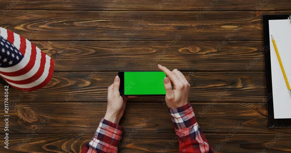 View from the top on the woman's hands scrolling and taping on the black smartphone with green screen on the wooden desk with American flag and blank paper sheet with a pencil. Horizontally. Chroma View from the top on the woman's hands scrolling and taping on the black smartphone with green screen on the wooden desk with American flag and blank paper sheet with a pencil. Horizontally. Chroma
