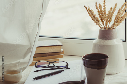 A stack of books, cup of coffee, bouquet of kernels, glasses, notebook and pen over a window. Classy attribute of intelligence reading learning. Moody. Fall mood. Autumn reading