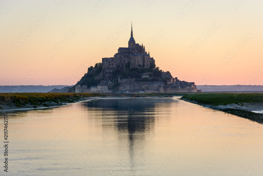 The silhouette of the famous Mont Saint-Michel tidal island in Normandy, France, at sunrise and high tide, reflecting in the still waters of the Couesnon river with the warm colors of the sky.