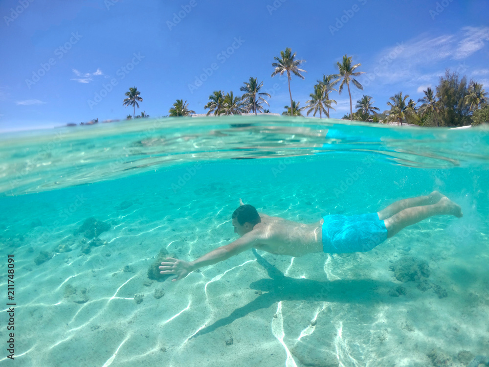 HALF UNDERWATER: man swimming along the white sand beach of tropical island.
