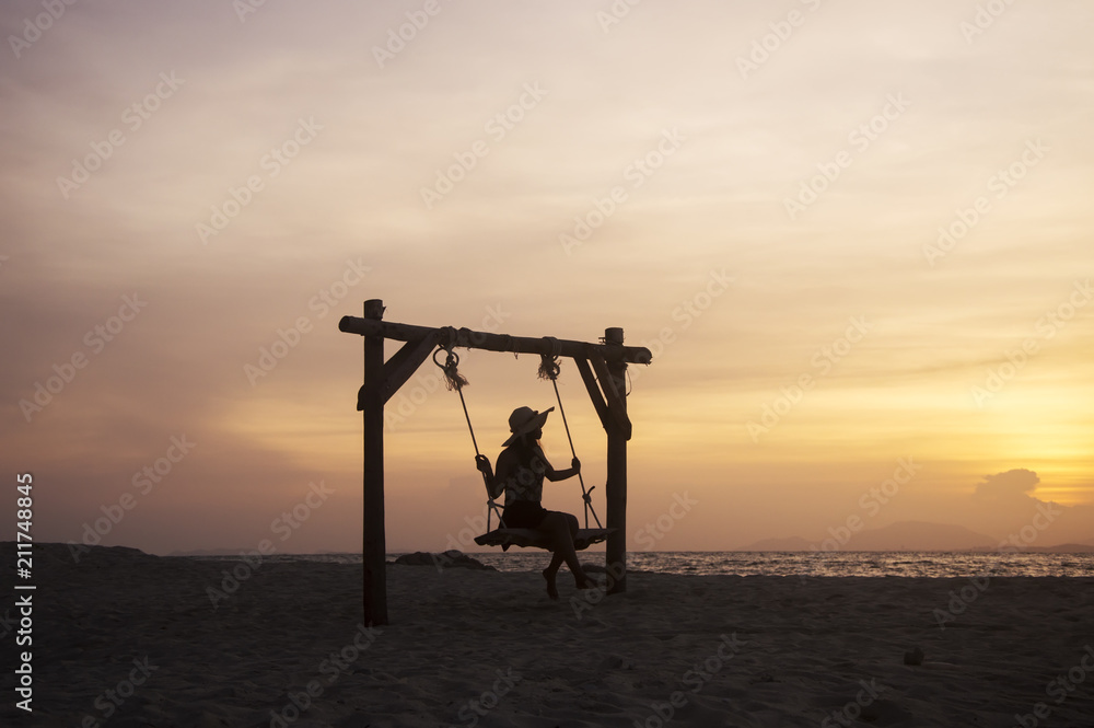 Fototapeta premium Young woman sitting on a swing at the beach with sunset sky
