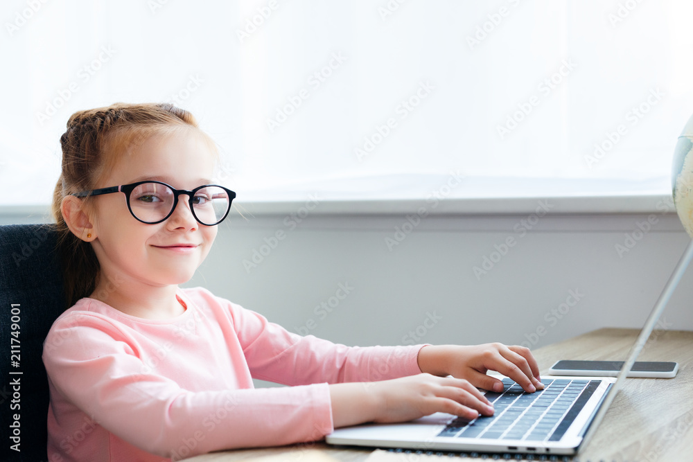 beautiful kid in eyeglasses using laptop and smiling at camera beautiful kid in eyeglasses using laptop and smiling at camera