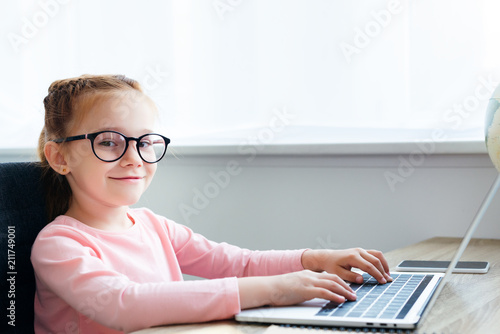 beautiful kid in eyeglasses using laptop and smiling at camera