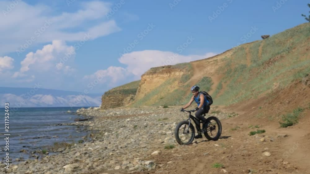 Fat bike also called fatbike or fat-tire bike in summer driving on the beach. The guy is riding heavily to the stony bank. The stones are big. The picture is contrast. Fat bike also called fatbike or fat-tire bike in summer driving on the beach. The guy is riding heavily to the stony bank. The stones are big. The picture is contrast.