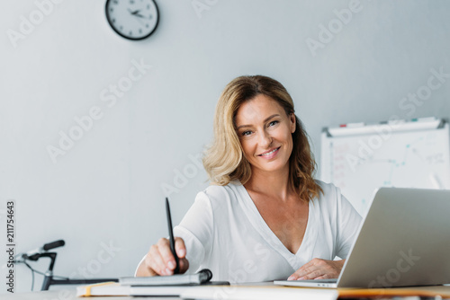 attractive businesswoman holding pen and looking at camera in office photo