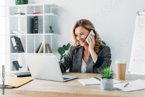 smiling attractive businesswoman talking by smartphone in office and looking at laptop