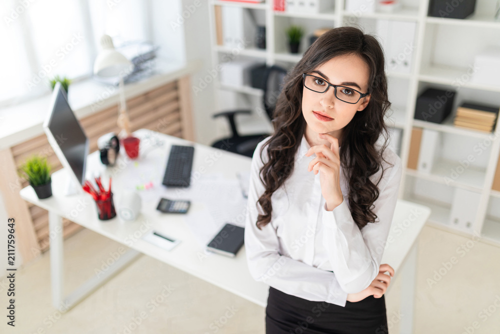 A beautiful young girl leans her hand on her chin and stands near the office table. A beautiful young girl leans her hand on her chin and stands near the office table.