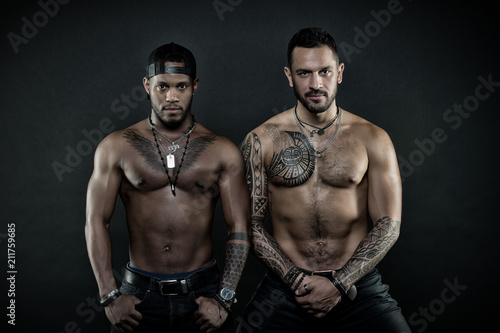 Machos with muscular tattooed torsos look attractive, dark background. Guys sportsmen with sexy muscular torsos. Athletes on confident faces with nude muscular chests. Sexy body concept