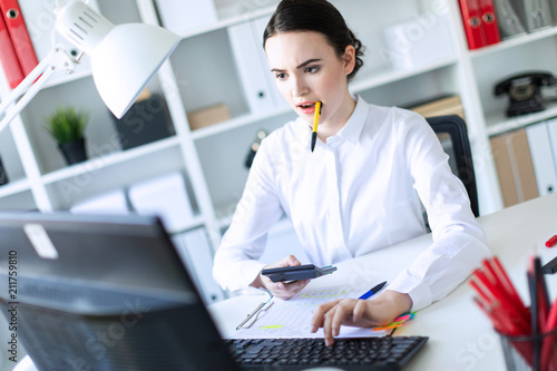 A young girl in the office holds a pen in her mouth and works with a calculator, documents and a computer. photo