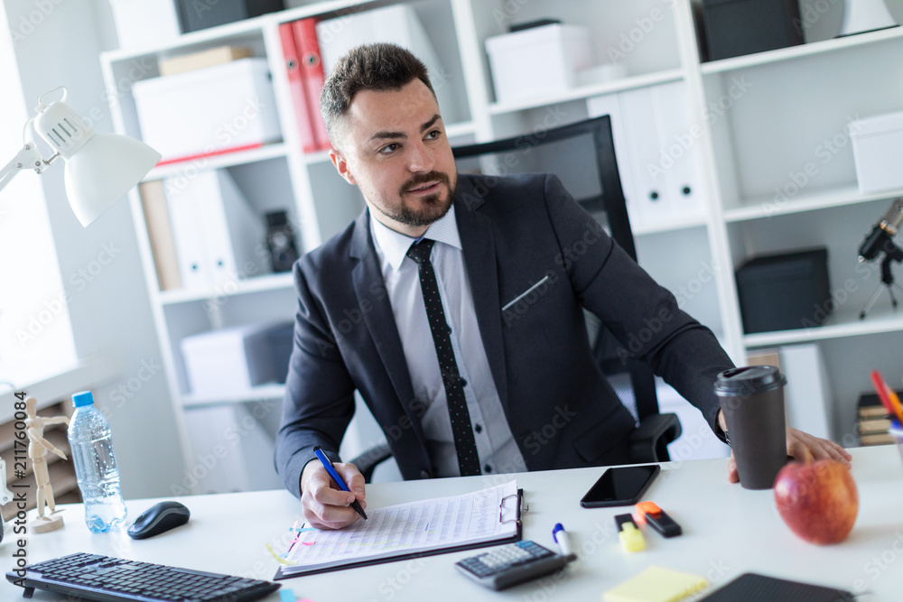 A man is sitting in the office at the table, holding a glass of coffee and a pen in his hand and working with documents. A man is sitting in the office at the table, holding a glass of coffee and a pen in his hand and working with documents.