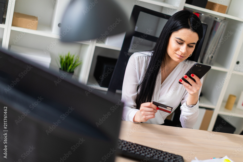 A young girl is sitting at the table, holding a bank card and phone. A young girl is sitting at the table, holding a bank card and phone.